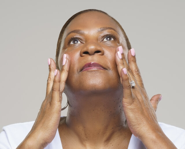 A person with short hair and a white shirt gently touching their face while looking upward against a light gray background.